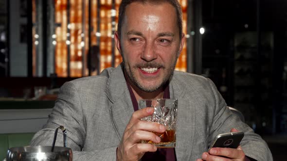 Handsome Mature Man Using Smart Phone While Tasting Whiskey at the Bar