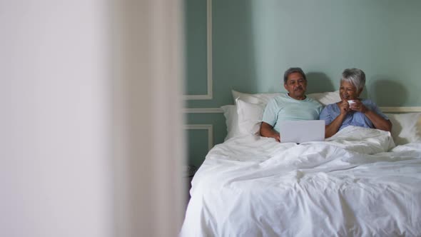 Happy senior mixed race couple sitting in bed using laptop