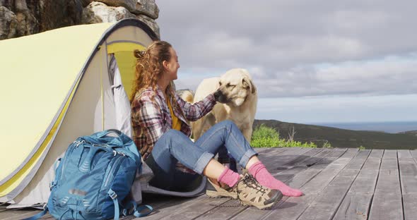 Happy caucasian woman camping sitting outside tent with pet dog, putting on boots on mountainside