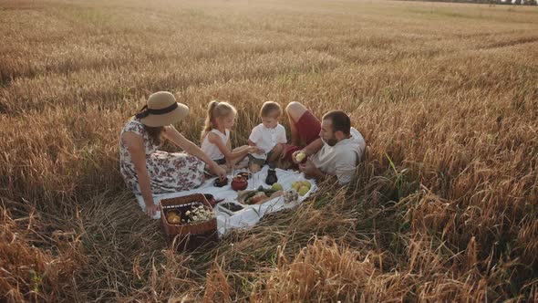 A Mom in a Straw Hat and a Father in Red Shorts Sit with Their Children at a Family Picnic in a