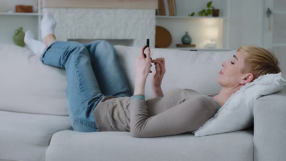 Relaxed Young Woman Lying on Sofa in Living Room Holding Smartphone Using Mobile Apps