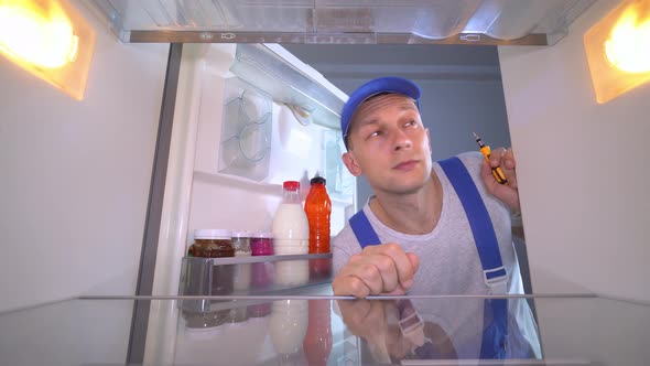 repairman opens door examines inside refrigerator with screwdriver in his hand.