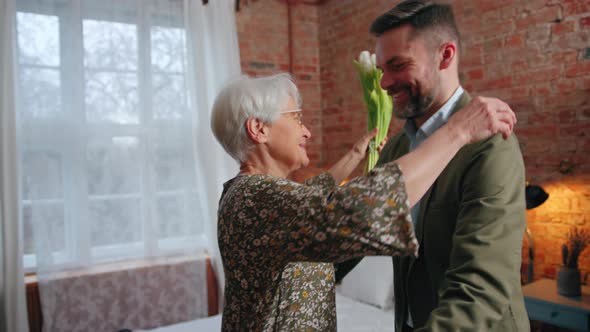 Cheerful Caucasian Grandmother Bonding with Her Adult Grandson Hugging and Holding Flowers