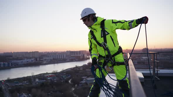 Industrial Climber in a Safety Harness Throws a Rope While Standing on the Edge of a Roof at Sunset