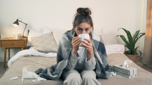 Sick Woman Covered with Blanket Holding Cup of Tea Sitting on Bed at Home