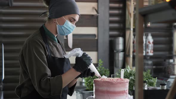 Female Baker Uses Precautions in a Medical Mask and Gloves Decorated Homemade Sweet Cake with Cream