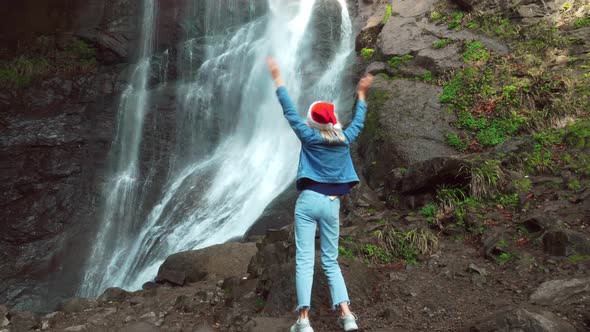 A girl in a Santa Claus hat and dances against the backdrop of a waterfall