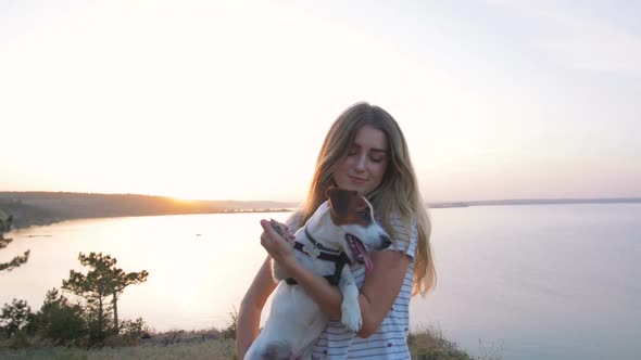Young Attractive Woman Playing with a Dog Jack Russell in the Meadow at Sunset with Sea Background