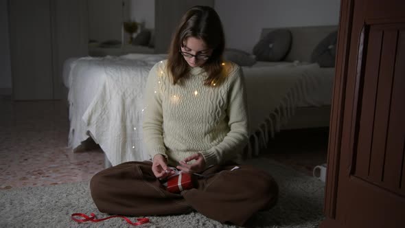 Girl with eyeglasses wearing beige sweater wrapping Christmas gift sitting on a capret near the bed