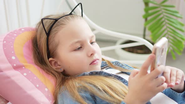 a Beautiful Little Girl in a Rim with Cat Ears with a Phone in Bed