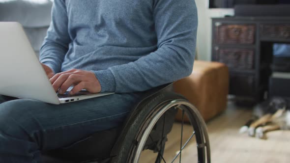 Midsection of caucasian disabled man in wheelchair using laptop in living room
