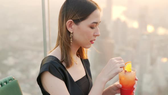 Asian woman drinking cocktail while waiting for friends at skyscraper rooftop restaurant at sunset.