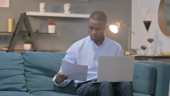 African Man with Laptop Having Success on Documents Sofa