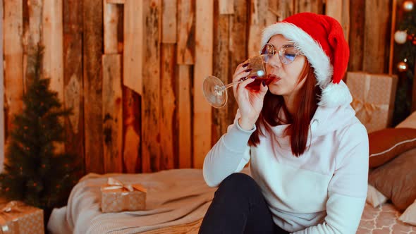 Young Woman in Santa Hat and Glasses Sitting on Bed and Drinking Red Wine