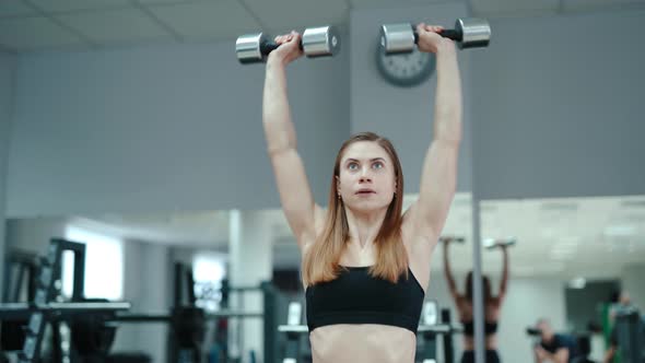 Pretty Woman in Black Top Raises Up Dumbbells on the Background of the Gym