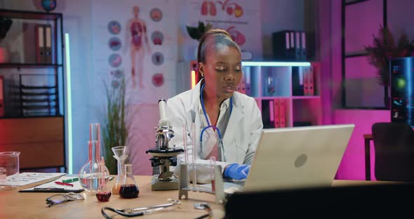 Female Chemist Sitting at Her Workplace in Lab with Different Test Tubes with Solutions