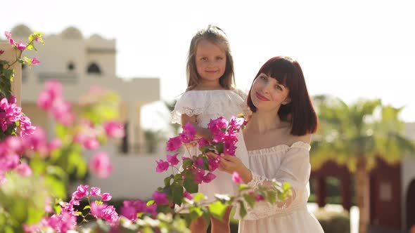 Smiling Mom with Daughter on White Dresses Sitting Against the Background of a Tropical Houses