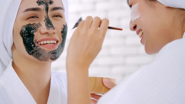 Beautiful young woman in white bathrobe applying a revitalizing black mask onto her friend's face.
