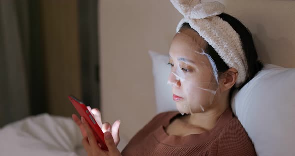 Woman use of mobile phone and apply the face mask on bed at night