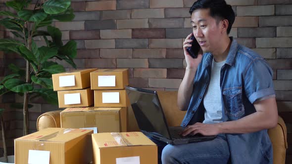 Asian Male Business Entrepreneur Using Laptop and Phone with Packs of Boxes at Home