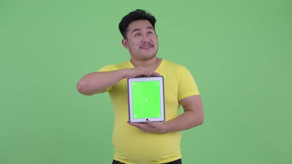 Happy Young Overweight Asian Man Thinking While Showing Digital Tablet