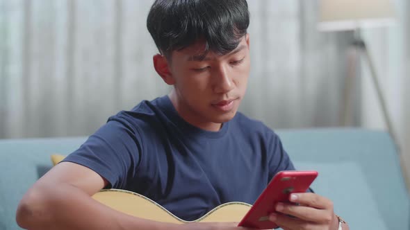 Close Up Of Asian Boy Composer With Note Book Looking At Smart Phone And Playing Guitar At Home