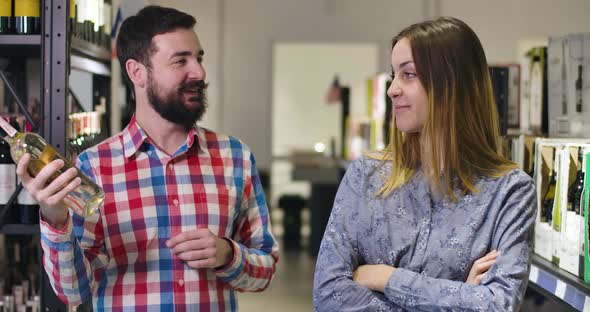 Sceptic Young Woman Looking at Cheerful Husband or Boyfriend Choosing Wine in Shop. Young Man and