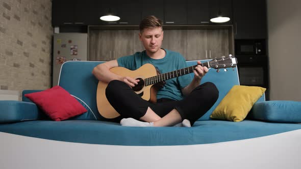 Caucasian Man Sitting on a Blue Sofa and Playing at the Guitar
