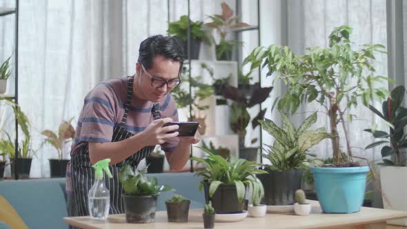 Asian Man Holding Smartphone And Taking Photos Of Plants At Home