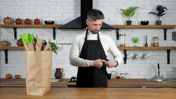 Bearded man chef in white sweater comes to home kitchen puts on black apron