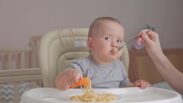 Mom feeds a newborn baby aged 12-17 months spaghetti with tomato sauce. Toddler eats pasta