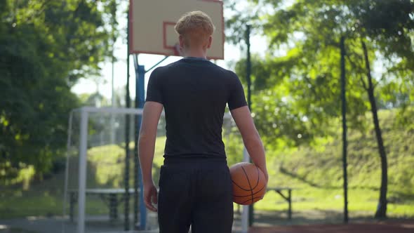 Camera Follows Redhead Sportsman Walking with Orange Ball To Basketball Hoop. Back View of Confident