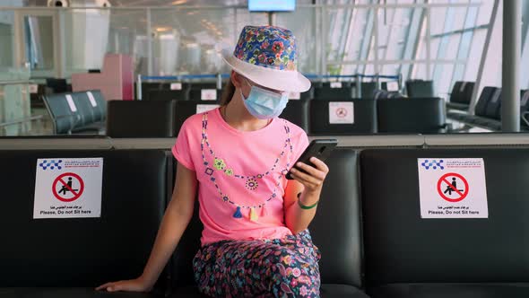 Teenage Girl, in Protective Mask, Uses Mobile. She Sitting in an Empty Airport Lounge, Waiting To