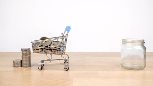 Stop motion animation shopping cart are carrying coins into a clear glass jar on wooden desk alt