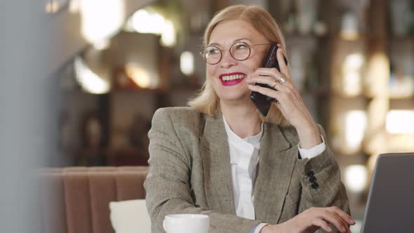 Senior Businesswoman Speaking on Phone and Using Laptop in Restaurant