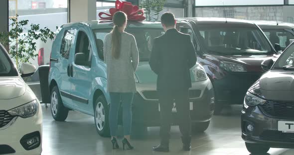 Wide Shot of Man and Woman Standing in Dealership in Front of Aquamarine Car. Back View of Trader