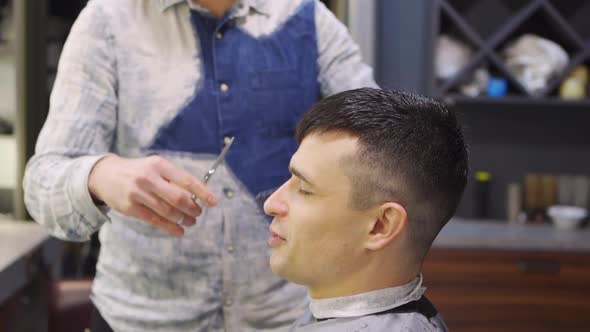 Hairdresser Makes a Haircut for a Brunet Man with a Scissors in a Barbershop