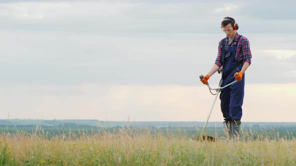 Young Man Mowing Grass with a Trimmer on a Picturesque Meadow