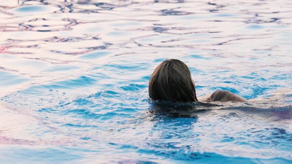 Woman Swims in a Pool with Clear Water on the Background of a Summer Sunset on Vacation