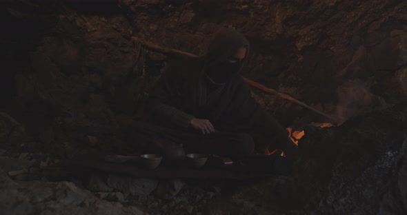 Hooded Figure In Cave With Campfire