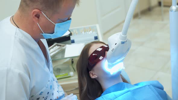A woman at the dentist in red UV protective glasses is undergoing laser teeth whitening. A dentist i