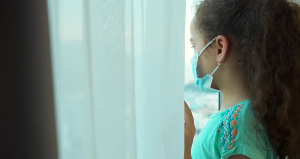 Portrait of a Cute Little Child or Little Girl in a Protective Medical Mask Looks Out the Window