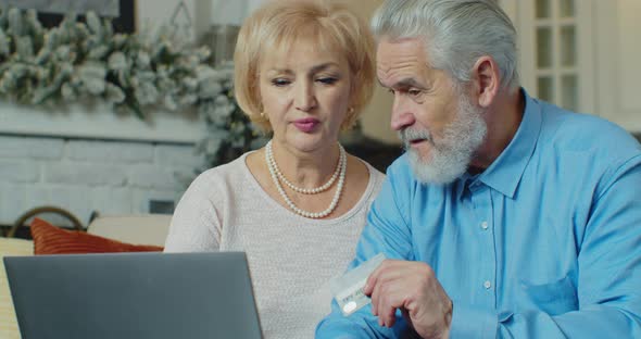 Medium Shot of Elderly Couple Talking While Making Online Payment Using Credit Card and Laptop
