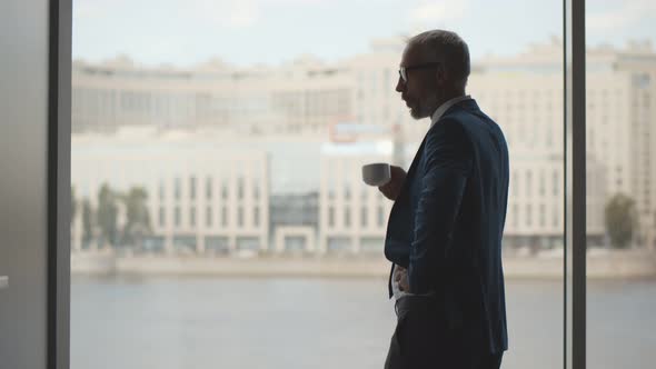 Happy Older Businessman Taking Break in Office Drinking Coffee Standing in Window