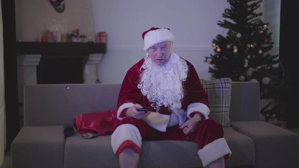 Old Man with False White Beard in the Costume of Santa Claus Switching Channels Sitting on the Sofa