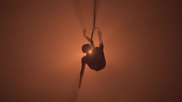 Silhouette of Flexible Aerial Gymnast Rotates on an Aerial Ring and Performs Acrobatic Tricks in the