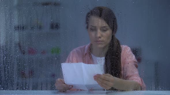 Upset Female Reading Letter Behind Rainy Window