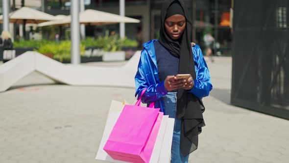 Black Shopaholic Adjusting Hijab and Using Smartphone