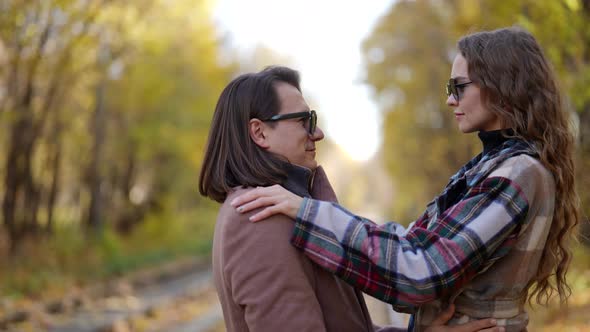 a Man and a Woman in a Coat and Sunglasses Hug Then Look at Each Other and Smile Against the