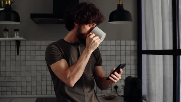 Closeup Portrait of Smiling Bearded Young Man Drinking Coffee From Cup Using Mobile Phone Watches